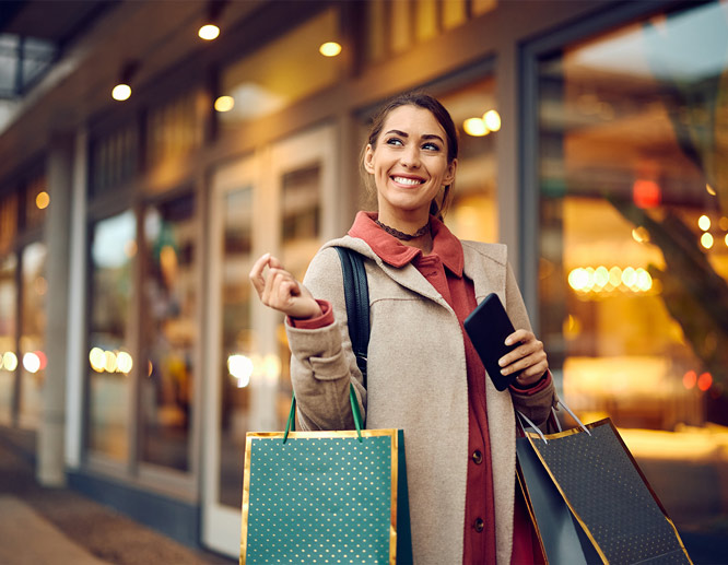 Smiling woman holding shopping bags outside boutique store, representing fall retail shopping and holiday promotions.
