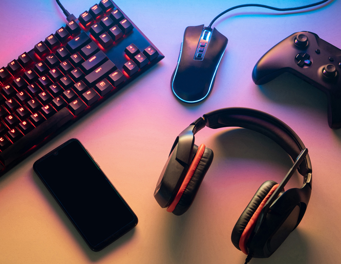 Gaming setup featuring a backlit keyboard, gaming mouse, headset, controller, and smartphone on a colorful lit background.
