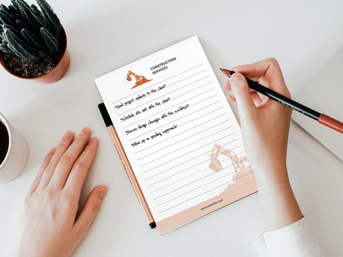 Construction-themed notepad printing with an excavator design, being written on with a cactus and coffee cup nearby