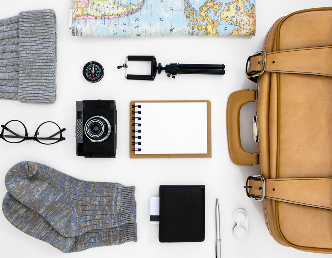 Flat lay of travel essentials including a map, camera, notebook, compass, glasses, socks, and a tan leather bag.